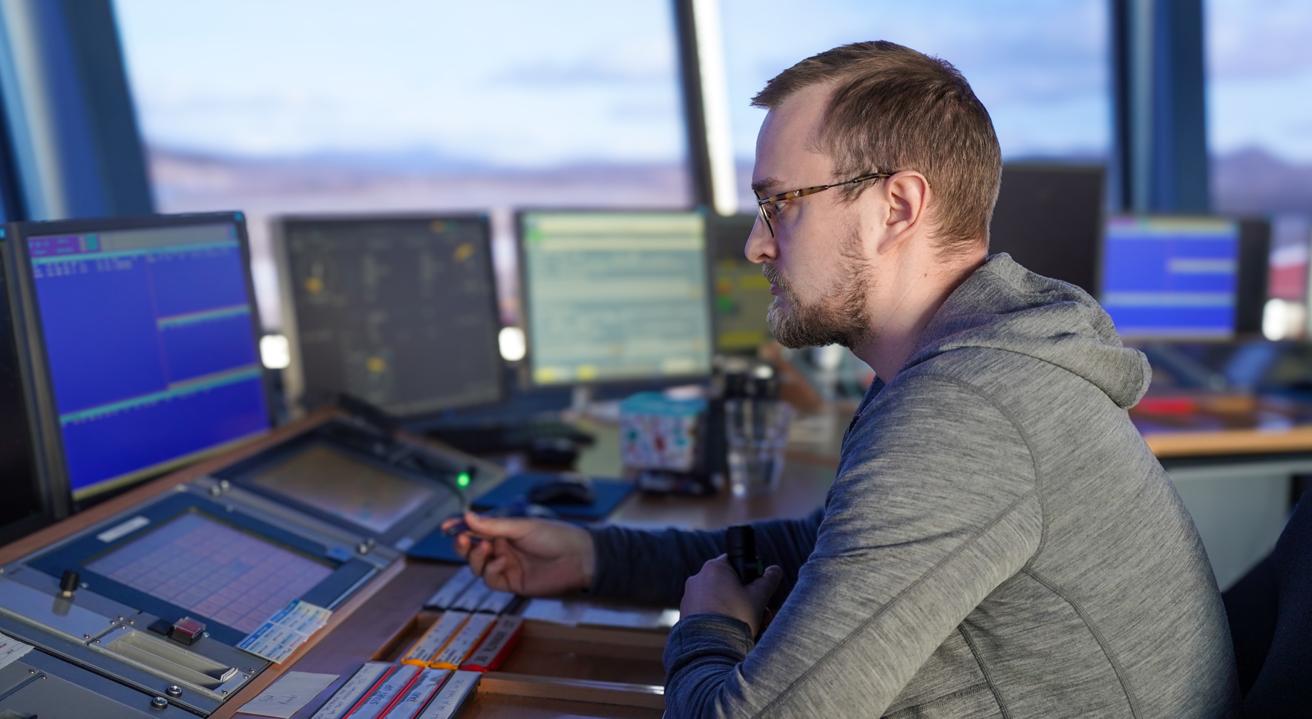 En mann i en kontrollrom omgivelse jobber med flere dataskjermer foran seg. Han har på seg en grå hettegenser og ser konsentrert ut. Skjermer viser informasjon som kan være relatert til overvåking eller kontrollfunksjoner.