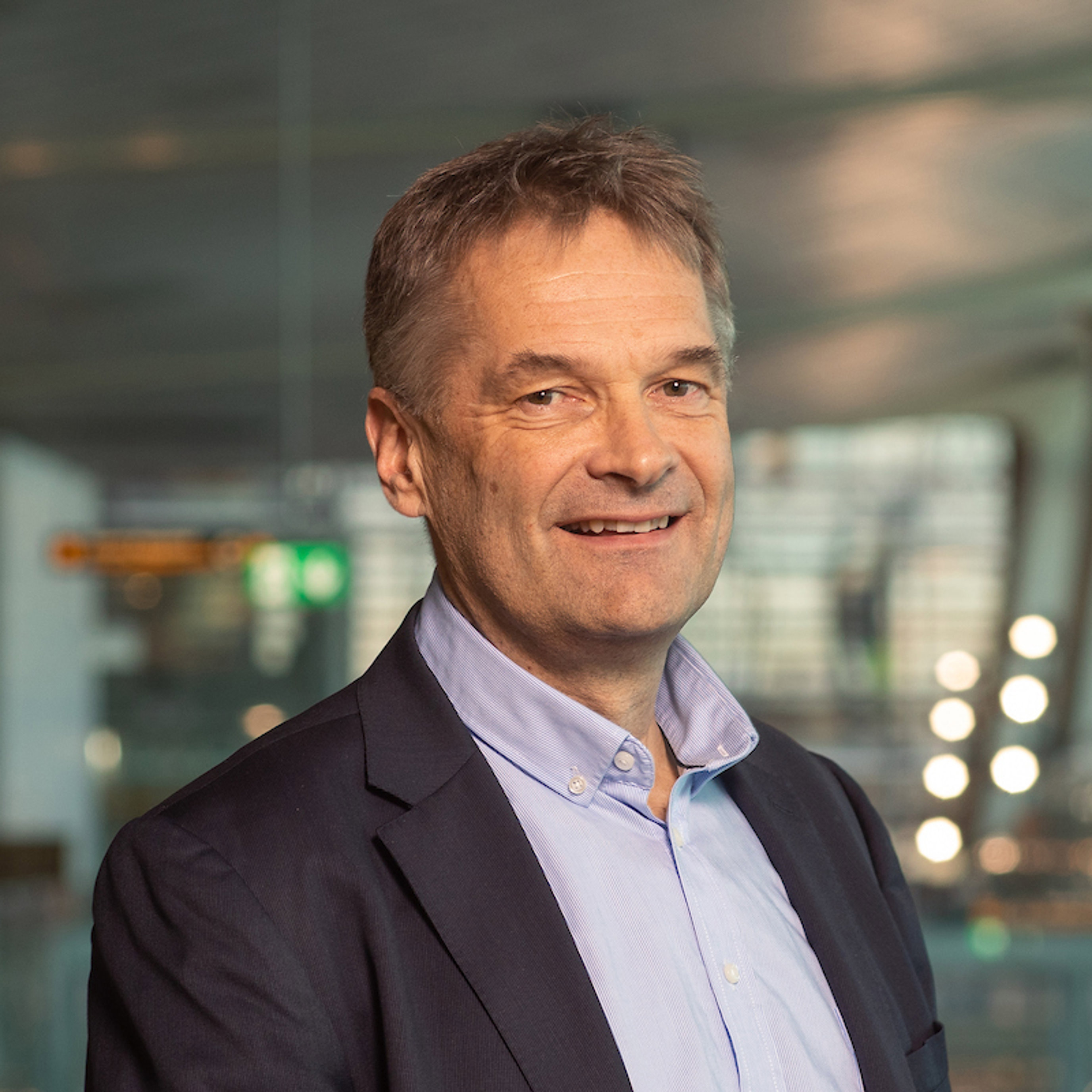 En person i dressjakke og skjorte, smilende i et moderne, profesjonelt innemiljø.