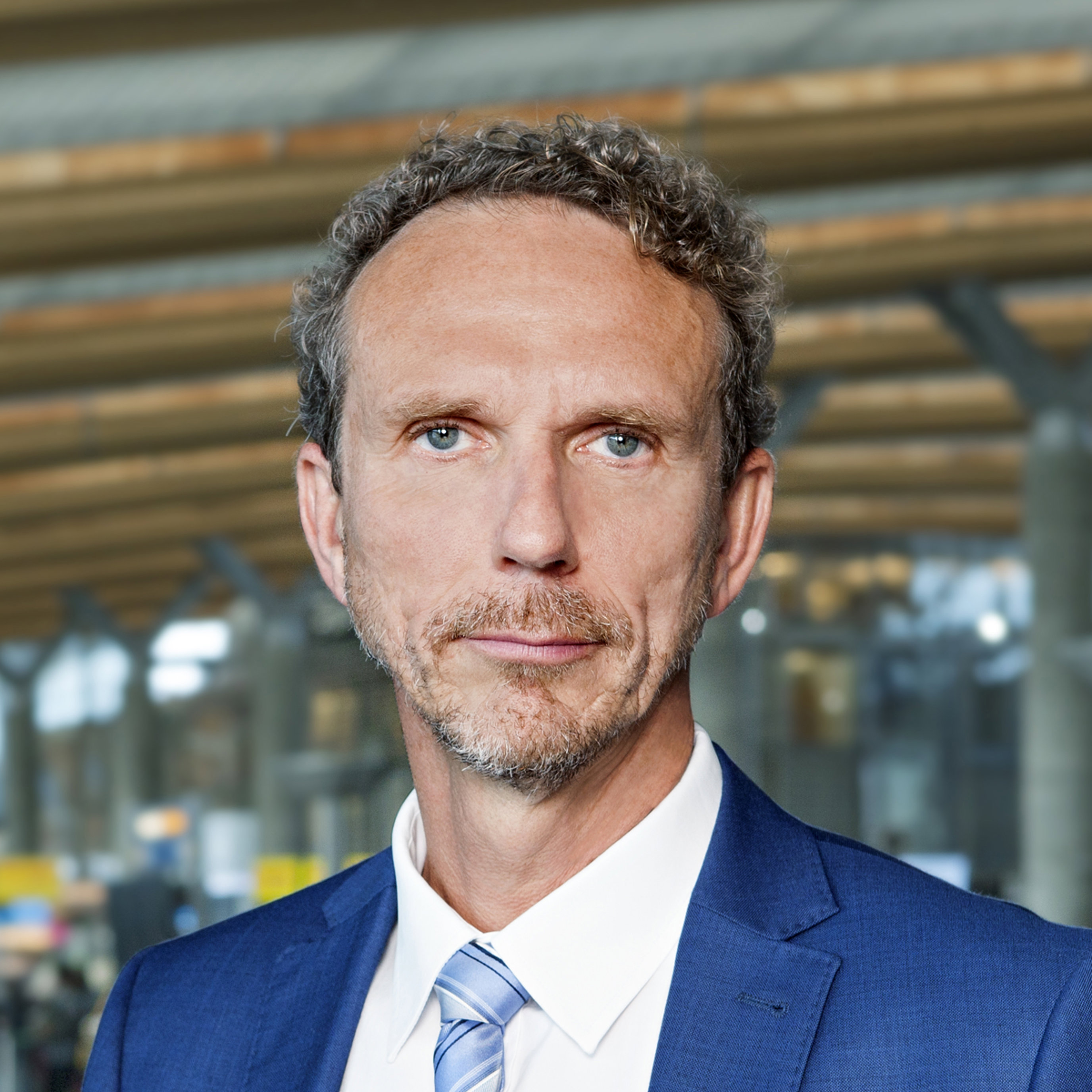 En mann i blå dress og slips står innendørs, sett mot en moderne, romslig bakgrunn med trepaneltak.