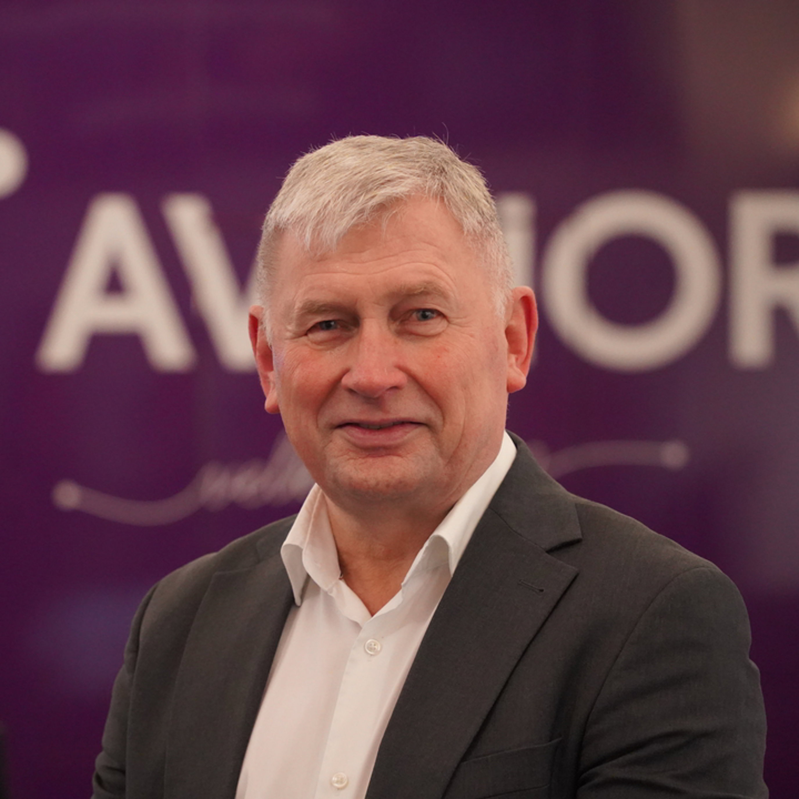 Mann med grått hår smiler foran en liten bakgrunn med teksten "AVINOR".