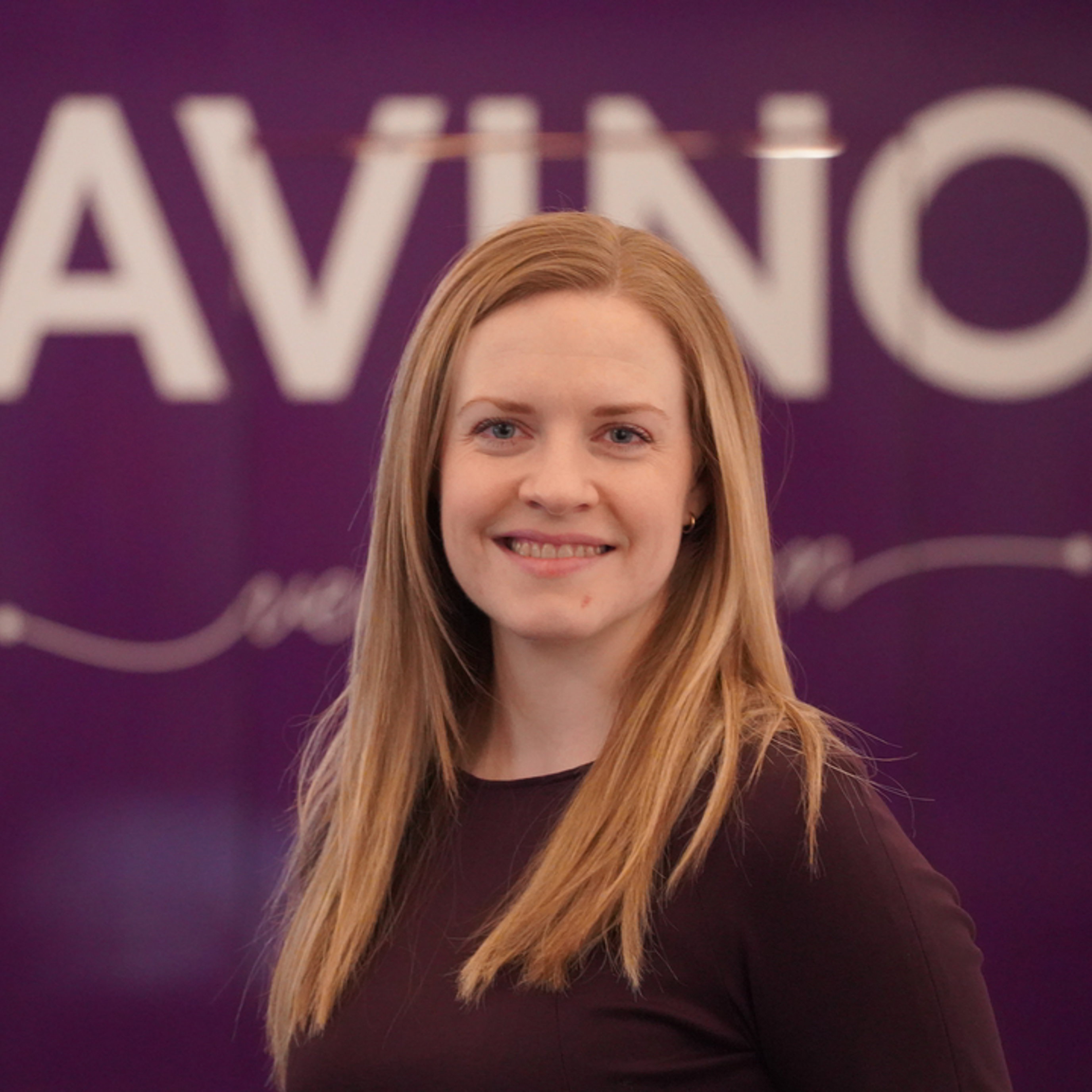 Kvinne med langt blondt hår smiler foran en lilla bakgrunn med teksten "AVINOR".