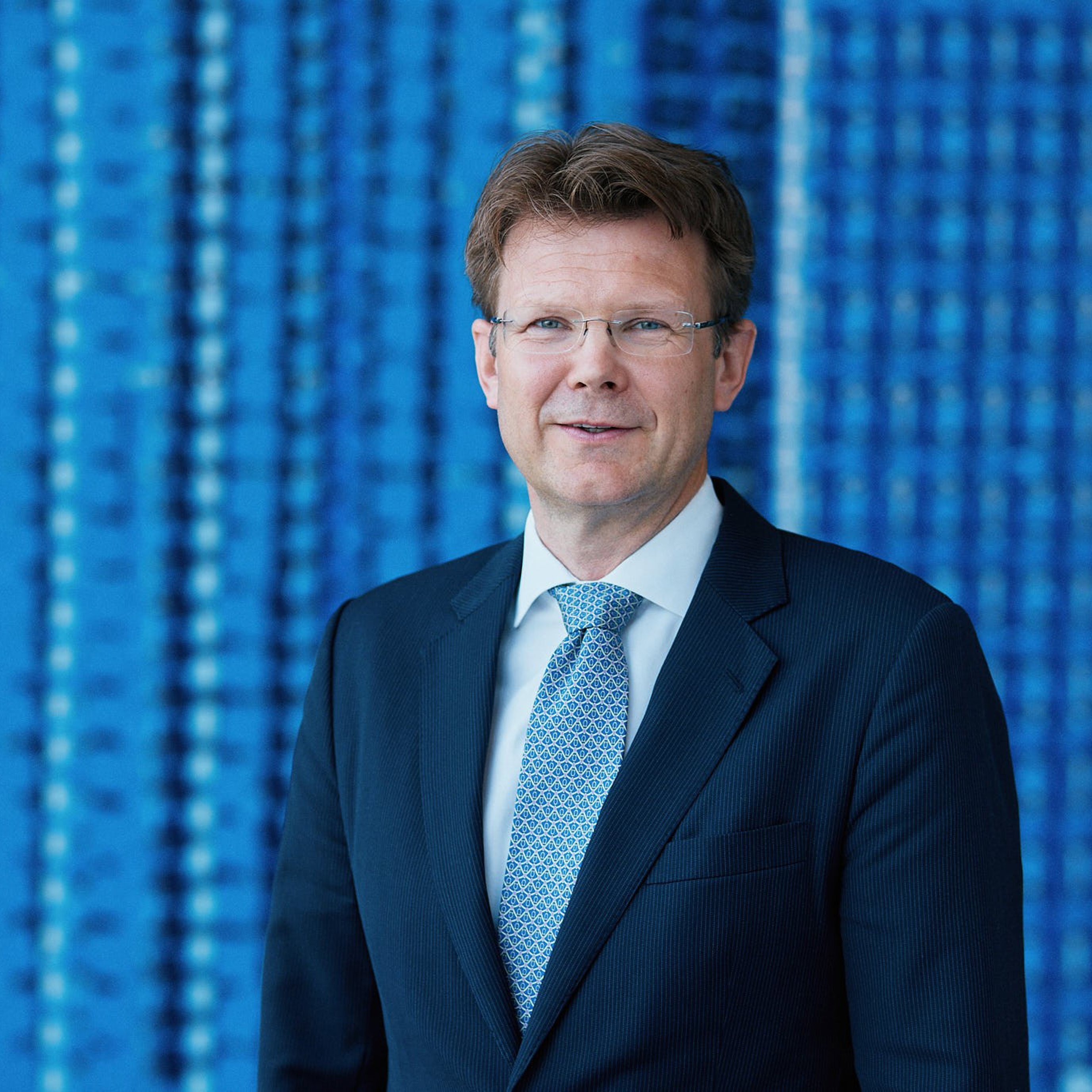 En mann i dress og slips står foran en blå bakgrunn med mønster.