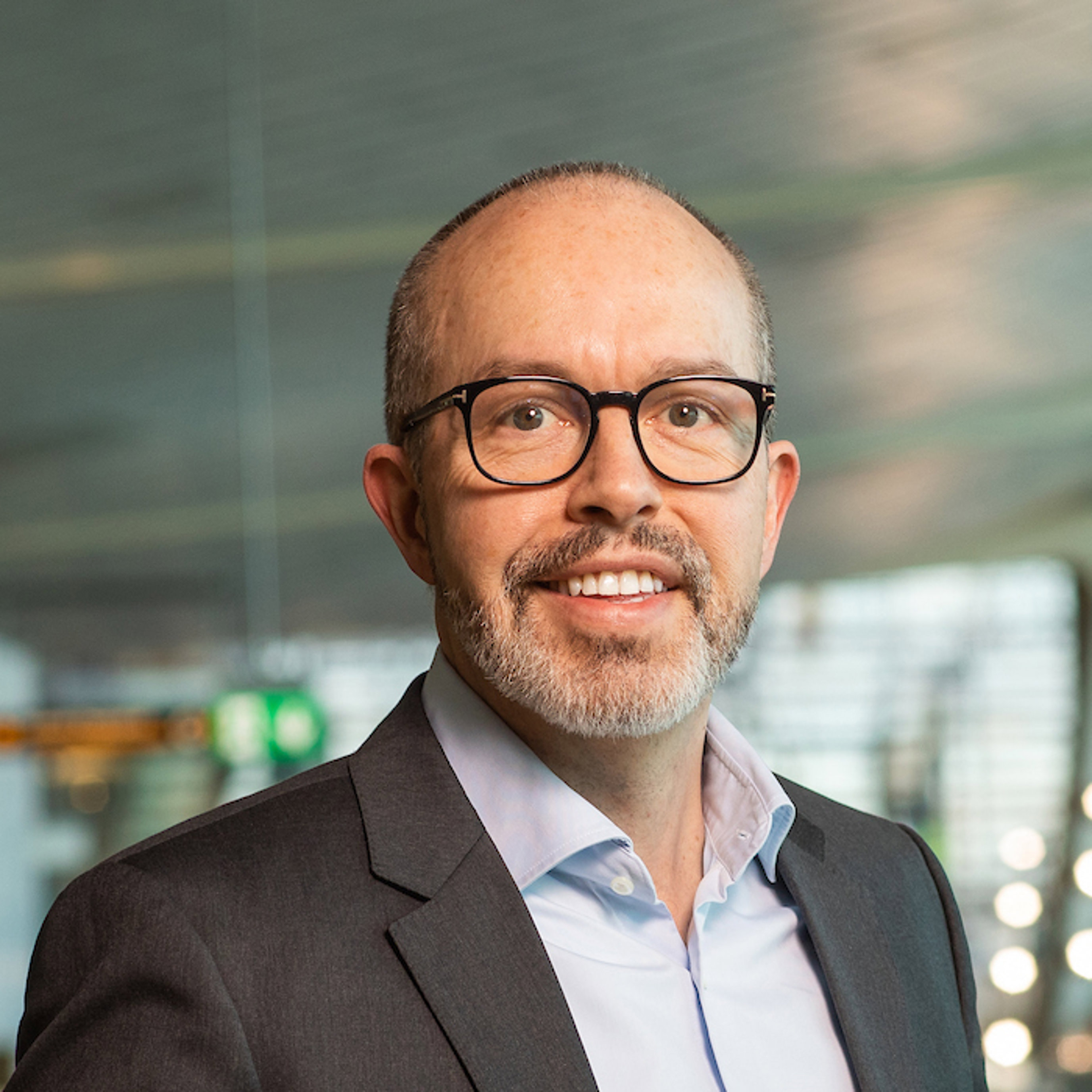 En mann med briller og skjegg i formell dressjakke smiler, med en modern kontorinnredning i bakgrunnen.