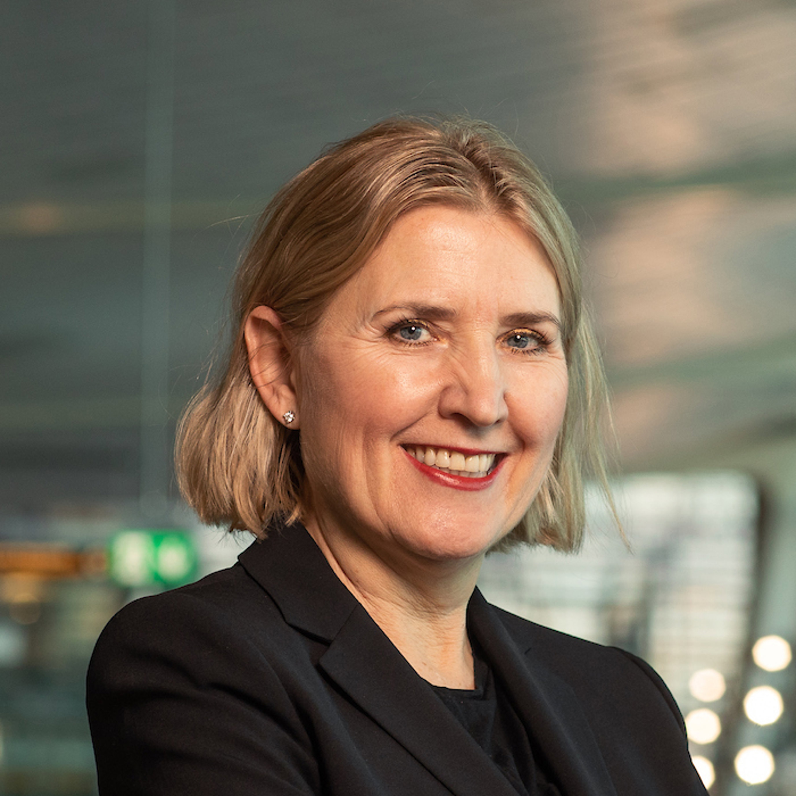 Kvinne med skulderlangt blondt hår og svart blazer smiler i et moderne kontormiljø.