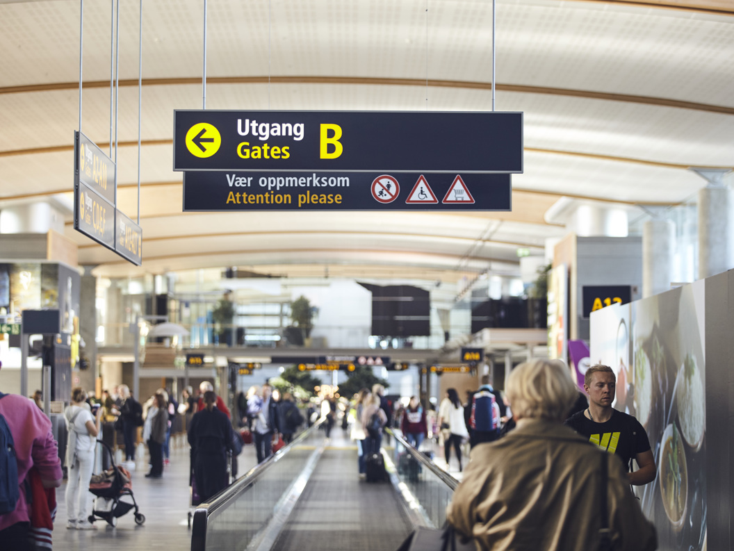 Folk på flyplass beveger seg mot utgang B, med skilt som viser vei og gir advarsler om røyking, mobilbruk, og rullestoltilgang.
