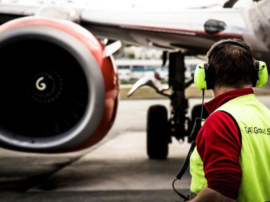 Flyplasspersonell med hørselvern overvåker et flys behandling på bakken foran en jetmotor.