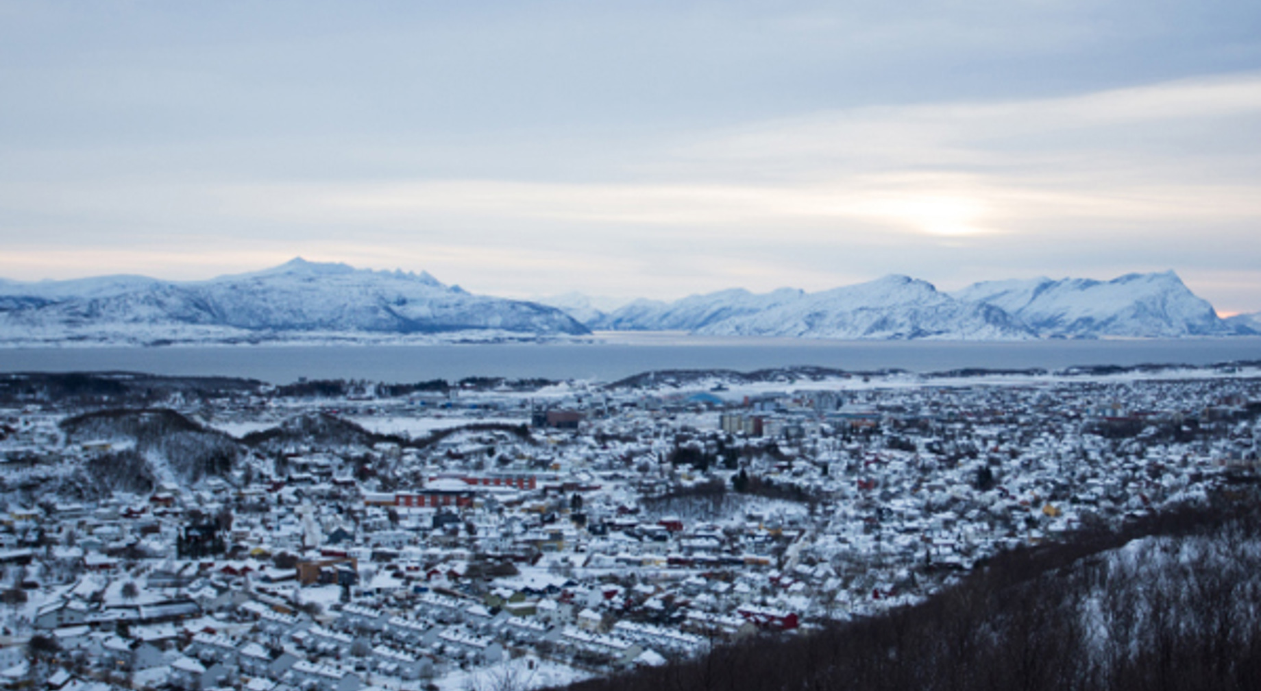 Vinterlandskap i Norge med snødekte fjell, hus, og en fjord i bakgrunnen under en overskyet himmel.