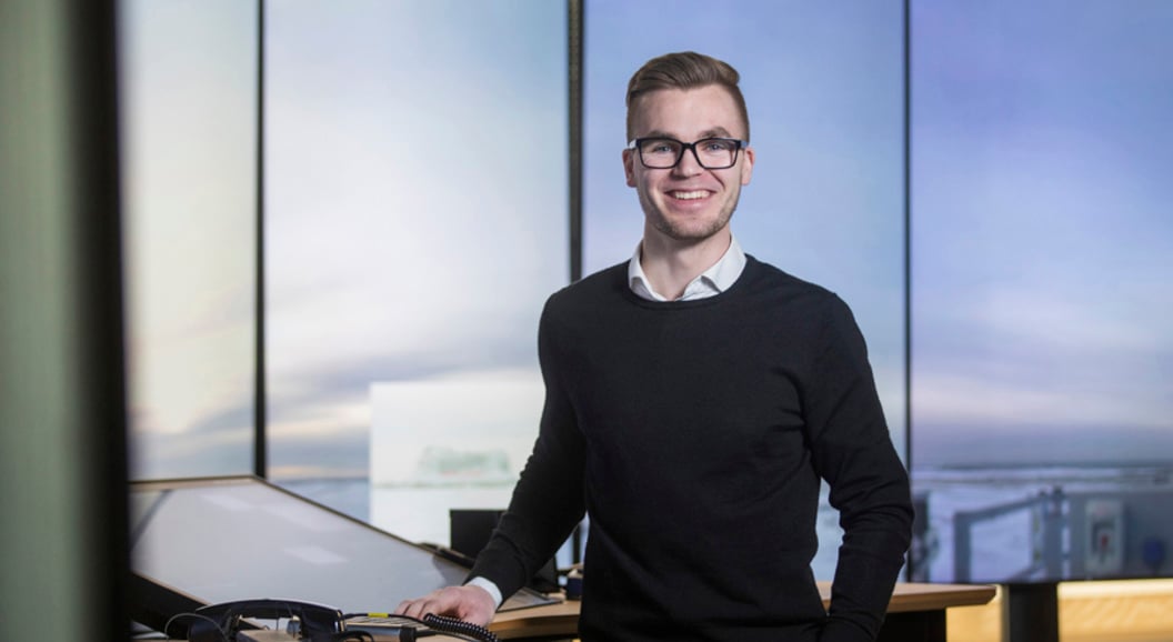 En smilende person med briller står ved et skrivebord med teknologiutstyr, mot en moderne kontorinnstilling.