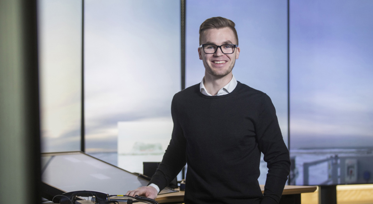 En smilende person med briller står ved et skrivebord med teknologiutstyr, mot en moderne kontorinnstilling.