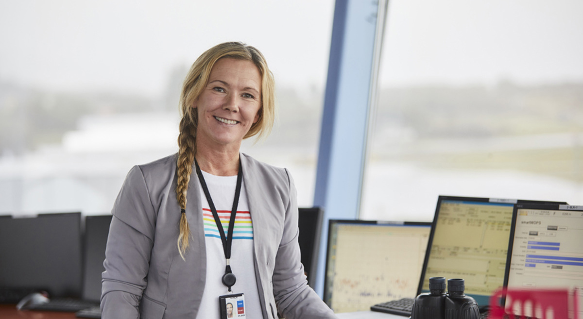 Kvinne i kontrollrom med dataskjermer i bakgrunnen, iført grå jakke og ID-kort.