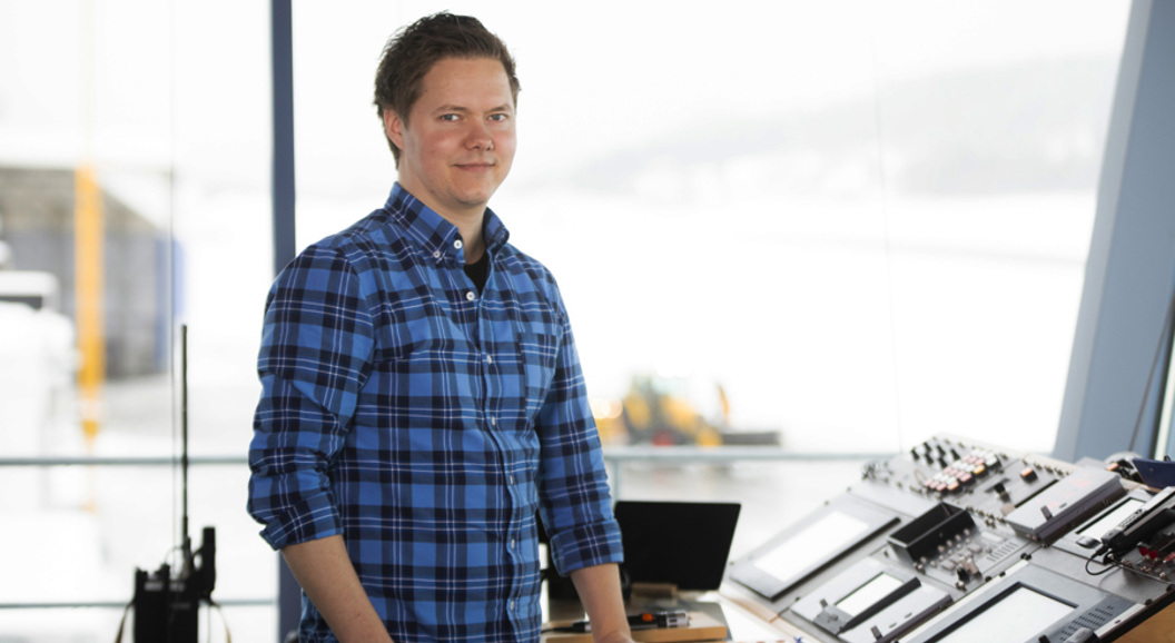 En person i blå rutete skjorte står ved et kontrollbord med skjermer og utstyr, med utsikt over en snødekt bakgrunn.