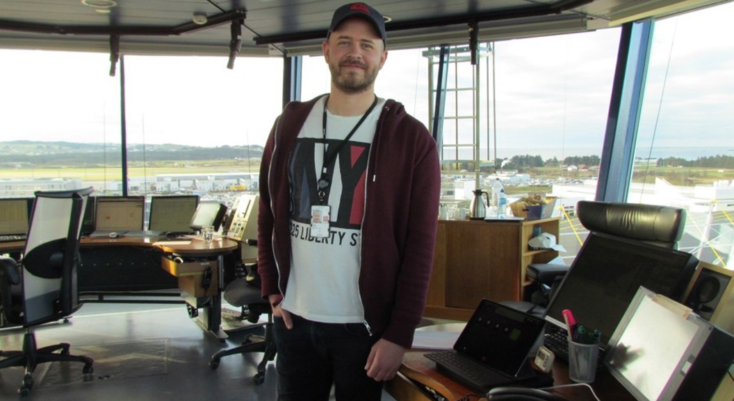 Mann i kontrolltårn med utsikt over flyplass, dataskjermer og utstyr synlig i bakgrunnen.