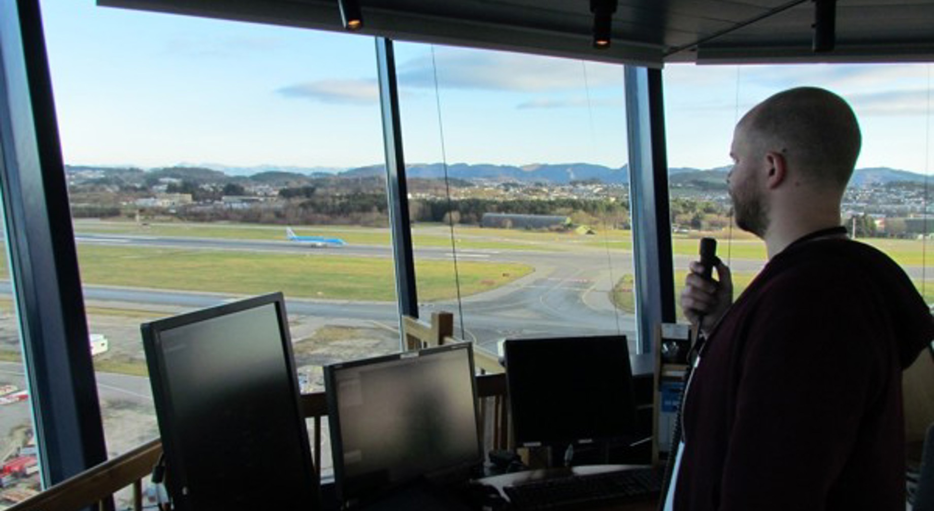 Flygeleder i kontrolltårn overvåker flytrafikk på flyplass, med utsikt over rullebanen og landskapet i bakgrunnen.
