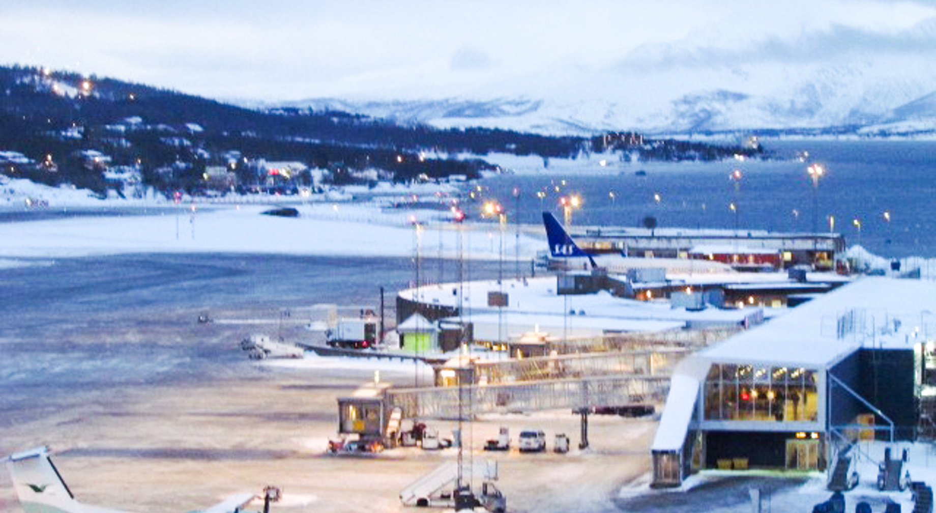 Lufthavn ved kysten i vinterlandskap med snødekte fjell i bakgrunnen, fly på bakken, og opplyste terminalbygg ved sjøen.