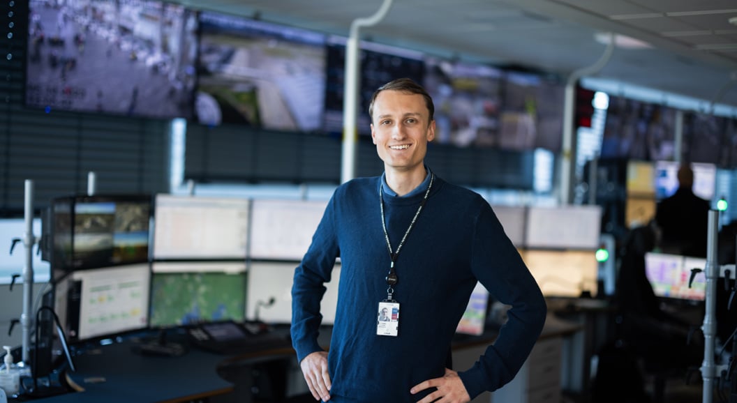 Mann i kontrollrom med flere skjermer, iført blå genser og ID-kort, smiler til kameraet. Bakgrunn viser en vegg med store overvåkingsskjermer.