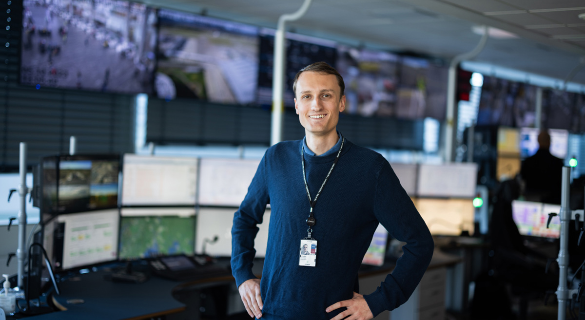 Mann i kontrollrom med flere skjermer, iført blå genser og ID-kort, smiler til kameraet. Bakgrunn viser en vegg med store overvåkingsskjermer.