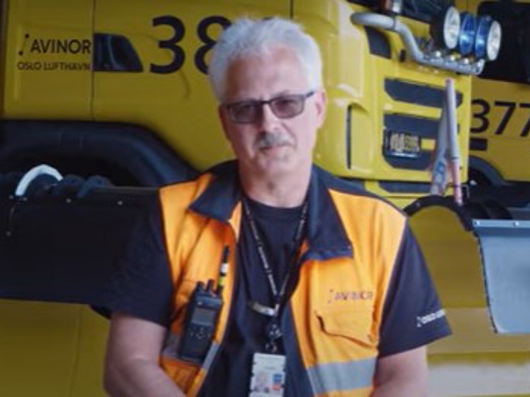 Mann i oransje vest foran gul snøryddemaskin fra Avinor, Oslo Lufthavn.