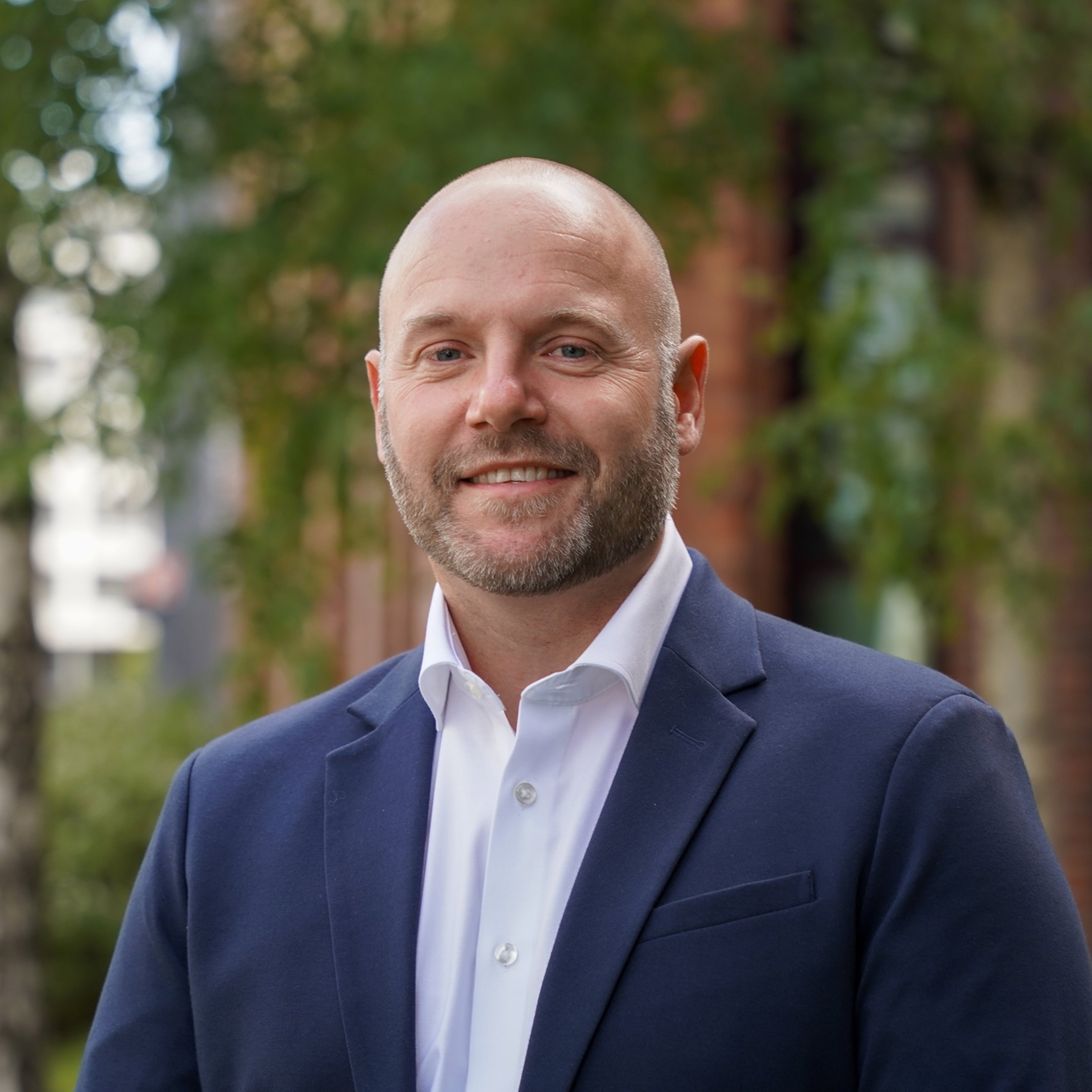 Person i formell blå dressjakke og hvit skjorte, smilende utendørs foran grønne trær og en rød murbygning.