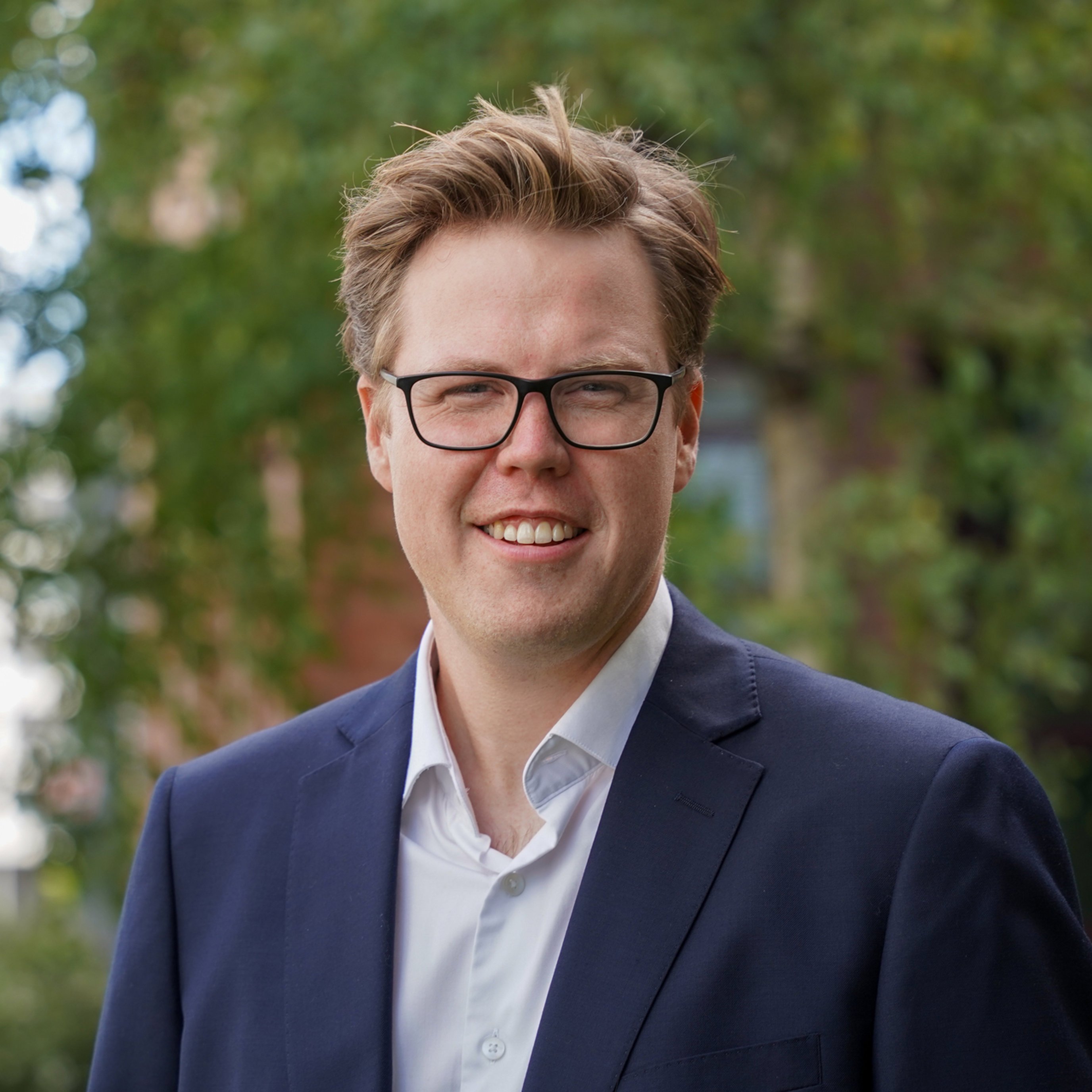 Mann med briller i dress smiler utendørs med grønt løvverk i bakgrunnen.