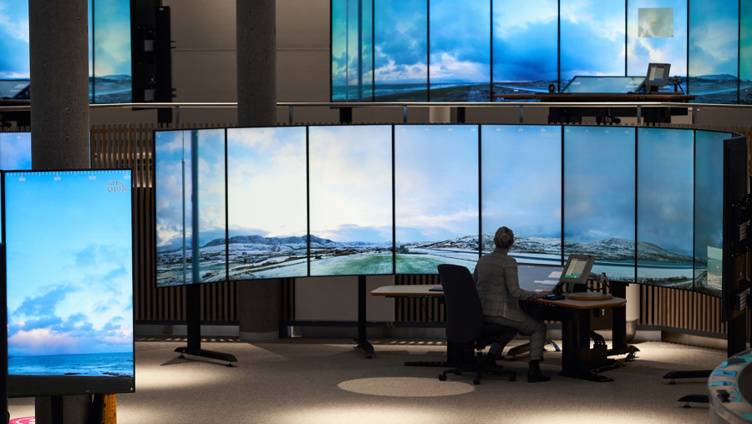 Person som sitter foran flere store skjermer på et kontrollsenter, med bilder av snødekte fjellandskap som vises på skjermene.