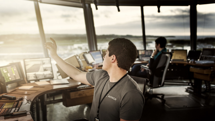 Mann i tårnkontroll med skjermer, peker ut av vinduet mens en kollega jobber i bakgrunnen på en flyplass, solnedgang i horisonten.