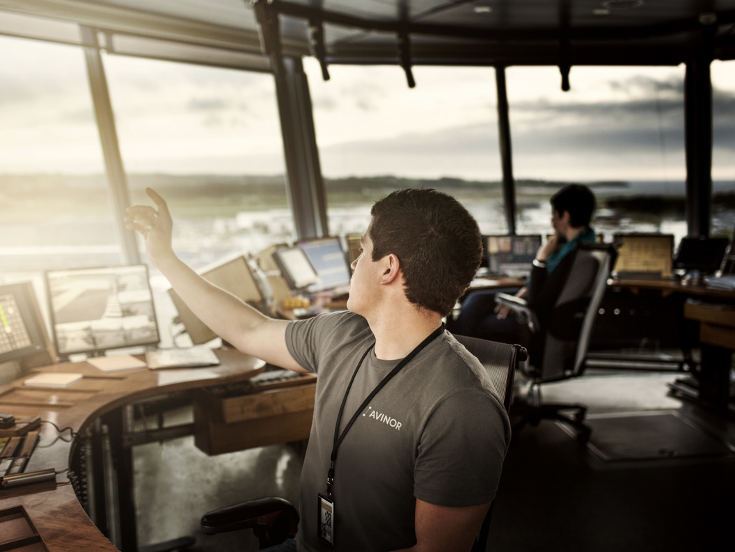 Mannlig flygeleder i et tårn, håndterer kommunikasjon med fly via konsoller, med utsikt til flyplassen i bakgrunnen.