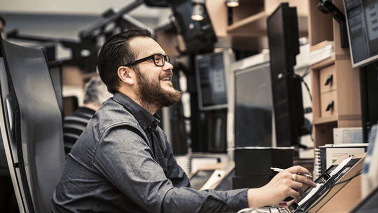 Mann med skjegg og briller smiler mens han arbeider på en datamaskin i et kontormiljø med flere skjermer.