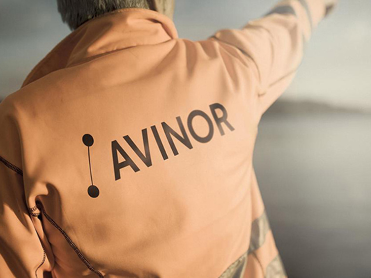 En mann med orange arbeidsjakke med Avinor logo på ryggen som peker oppover mot skyene, fokus på logoen.
