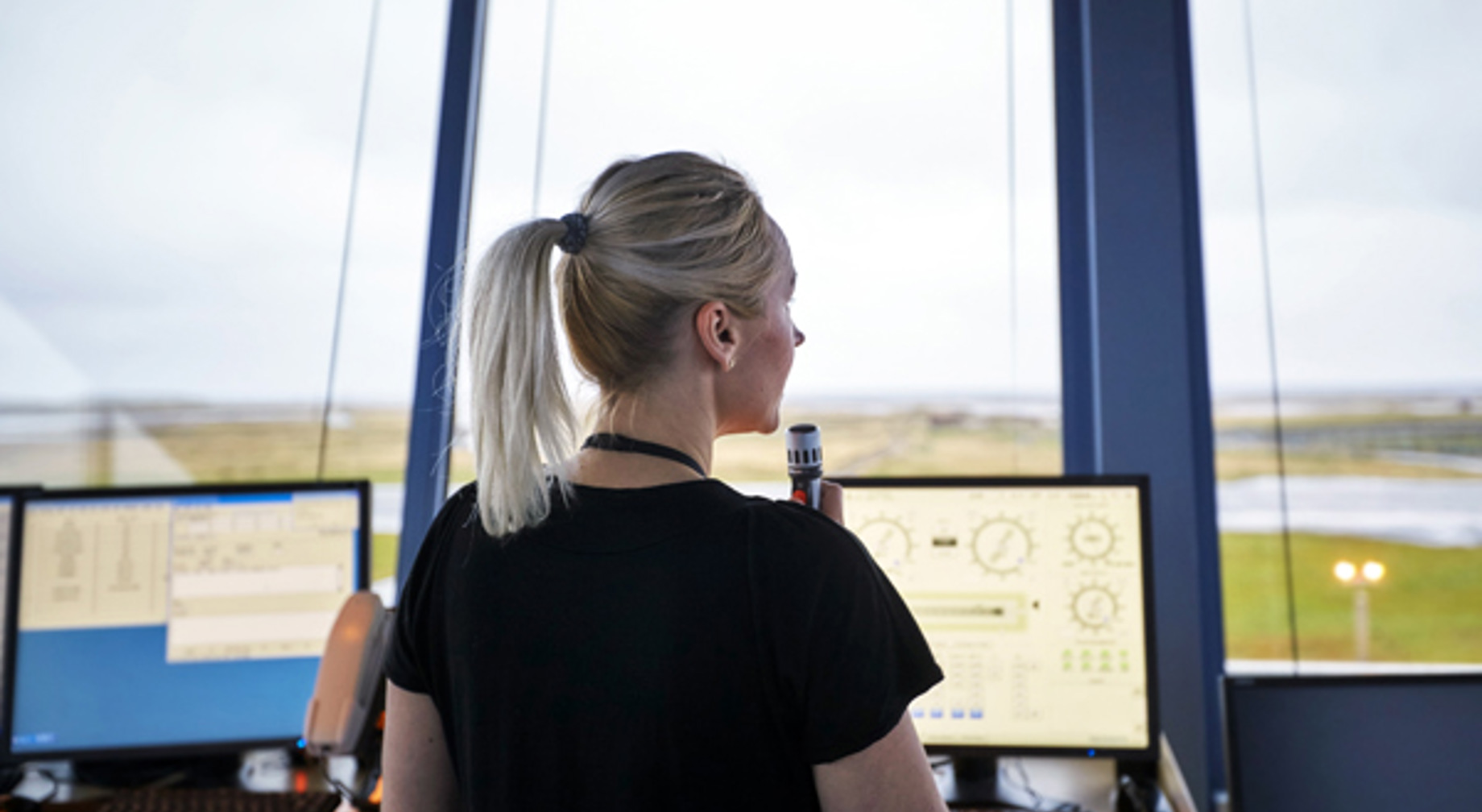 Lufttrafikkontrollør i tårn med ryggen til mens hun kommuniserer via radio, ser ut av vinduet mot rullebanen. To dataskjermer viser lufttrafikkdata.