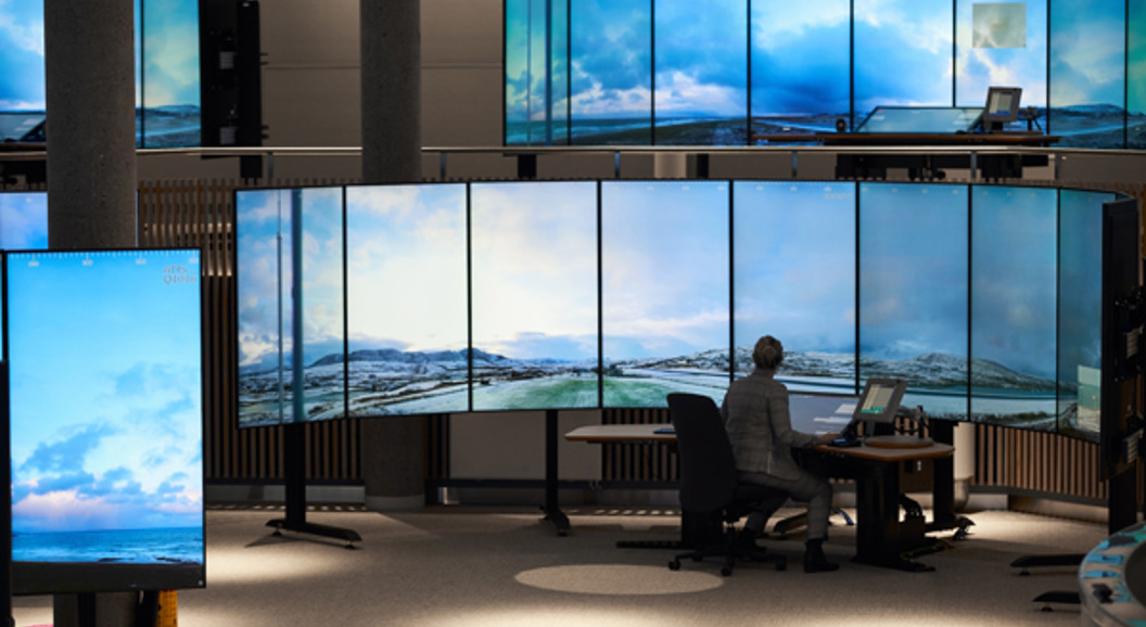 En person sitter ved et kontrollrom omgitt av store skjermer som viser et landskap av himmel og fjell.