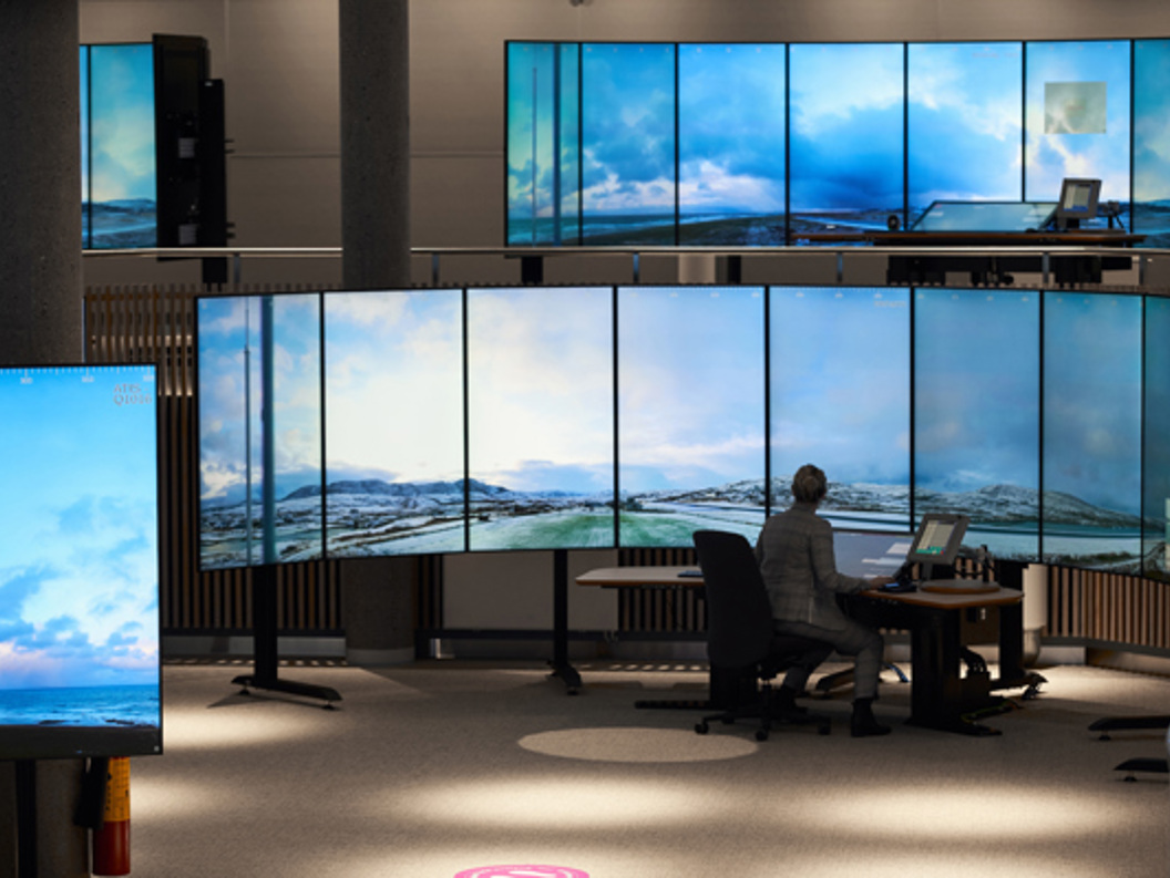 En person sitter ved et kontrollrom omgitt av store skjermer som viser et landskap av himmel og fjell.